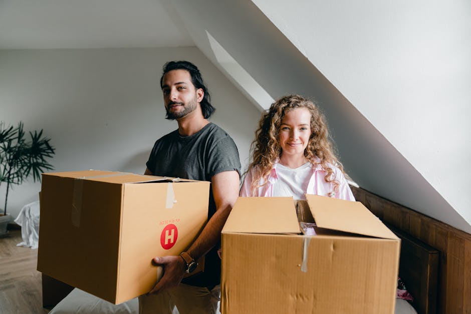 A man and a woman stand inside a residential property, each holding a large cardboard box prepared for a home relocation. The man, with dark hair and a beard, is wearing a grey t-shirt and holds a sealed cardboard box marked with a red 'H' label, positioned in front of him. The woman, with curly blonde hair, is dressed in a light-colored top and holds an open box with packing paper, ready for moving. They are next to a wooden headboard against a white wall with a sloped ceiling and natural daylight illuminating the space. In the background, there is a potted plant on a stand and a white covered object, possibly furniture or decorative items, indicating an indoor environment during packing and furniture transport preparations. This image is associated with house removals services, such as those offered by Hornchurch Man and Van, involving the packing, loading, and transportation process involved in home moving and furniture transport.