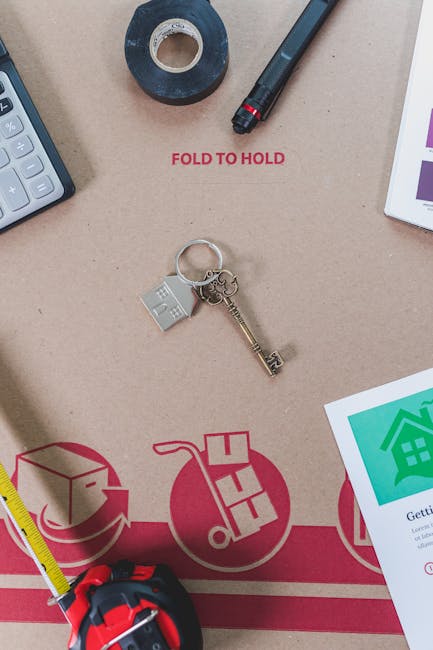 A flat-lay image of various items related to house removals and packing placed on a beige surface. This includes a black marker pen, a roll of black adhesive tape, and a white calculator partially visible on the left side. In the centre, there is a set of keys attached to a keyring with a small silver house-shaped charm, resting on a cardboard box marked with red illustrations of a house and packing symbols, along with texts like 'FOLD TO HOLD'. To the right, part of a white tablet displaying a green house icon and a document with a colourful chart is visible. The bottom left shows a yellow retractable measuring tape, and near the top, the phrase 'FOLD TO HOLD' is printed in red. These objects suggest a home relocation process involving packing, planning, and preparation for moving, consistent with services offered by Hornchurch Man and Van.