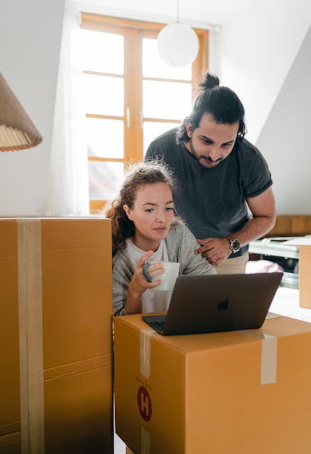 A man and a woman stand inside a residential property, each holding a large cardboard box prepared for a home relocation. The man, with dark hair and a beard, is wearing a grey t-shirt and holds a sealed cardboard box marked with a red 'H' label, positioned in front of him. The woman, with curly blonde hair, is dressed in a light-colored top and holds an open box with packing paper, ready for moving. They are next to a wooden headboard against a white wall with a sloped ceiling and natural daylight illuminating the space. In the background, there is a potted plant on a stand and a white covered object, possibly furniture or decorative items, indicating an indoor environment during packing and furniture transport preparations. This image is associated with house removals services, such as those offered by Hornchurch Man and Van, involving the packing, loading, and transportation process involved in home moving and furniture transport.