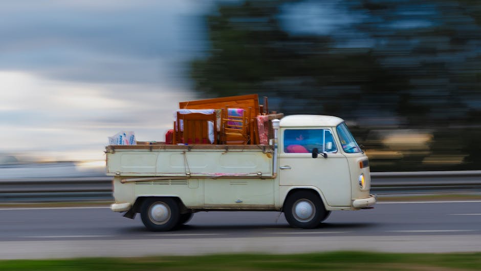 A vintage cream-coloured van is travelling along a road during daytime, with a blurred background indicating motion. The van's cargo area is loaded with various furniture items, including a wooden chair, small tables, and wrapped items, all secured for transportation. The interior of the van can be partially seen through the front windshield, which has a pink object visible inside. The scene captures a home relocation or furniture transport process, with the van from Hornchurch Man and Van engaged in a professional removals operation. The background, featuring trees and sky, is blurred to emphasize the movement and loading process of furniture during a house moving service, illustrating logistics involved in packing and transporting household items efficiently.