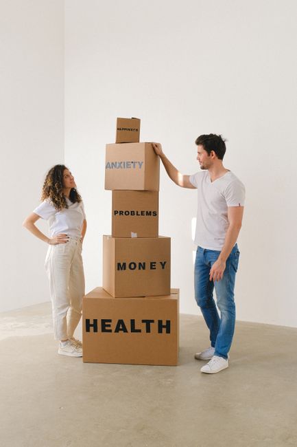 A young man and woman are standing indoors on a light-colored floor against a plain white wall, engaging in a symbolic stacking of large cardboard boxes. The woman, dressed in a white T-shirt and cream-colored trousers, stands with her hands on her hips and looks up at the boxes. The man, wearing a white T-shirt and blue jeans, is placing the top box gently on the stack. The boxes, which are arranged vertically, have labels in bold black and blue letters reading 'HEALTH,' 'MONEY,' 'PROBLEMS,' 'ANXIETY,' and the top box labeled 'HAPPINESS.' Surrounding the boxes are visible packing materials, although not detailed, emphasizing a moving or home relocation theme associated with furniture transport and packing. The scene likely represents themes of moving logistics, packing, and mental well-being during house removals, as featured on hornchurchmanandvan.com’s services page for bulky item removals, access solutions, and house relocations.