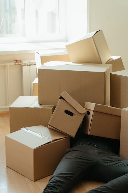 A person lying on the floor amidst a collection of cardboard boxes of various sizes, some sealed and others open, during a house move in a bright room with a large window and radiator visible in the background. The boxes, used for packing belongings, are stacked and scattered around, with a cardboard box featuring built-in handles leaning against another. The scene depicts a packing and moving process associated with home relocation services, likely arranged by Hornchurch Man and Van, as part of their end of tenancy removals in Hornchurch. The room is well-lit with natural light, and the person appears to be in the midst of handling or organizing the packed items, ready for transportation by van for a smooth furniture transport or home relocation.