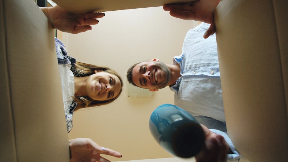 A group of two men and one woman are engaged in a home relocation process, seen from inside a cardboard box with an open top, looking downward into the camera. The scene occurs inside a well-lit room with neutral-colored walls. The woman, on the left side, has long brown hair and is smiling, wearing a checkered shirt. The man in the center has short hair, a beard, and is also smiling, dressed in a light blue button-up shirt. The third person, partially visible at the bottom right, is holding a black cylindrical object, possibly a parcel or packaging material. They are surrounded by packaging items such as cardboard, plastic wrap, and packing blankets, indicating an active packing and moving process facilitated by Hornchurch Man and Van. This setup exemplifies furniture transport and packing procedures during end of tenancy removals, with the focus on careful handling and organized logistics before loading items into a moving van. The company's services support efficient house removals and home relocation tasks, as shown in this orderly packing scene.