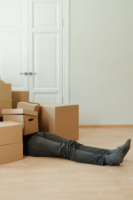 A person lying on the floor amidst a collection of cardboard boxes of various sizes, some sealed and others open, during a house move in a bright room with a large window and radiator visible in the background. The boxes, used for packing belongings, are stacked and scattered around, with a cardboard box featuring built-in handles leaning against another. The scene depicts a packing and moving process associated with home relocation services, likely arranged by Hornchurch Man and Van, as part of their end of tenancy removals in Hornchurch. The room is well-lit with natural light, and the person appears to be in the midst of handling or organizing the packed items, ready for transportation by van for a smooth furniture transport or home relocation.