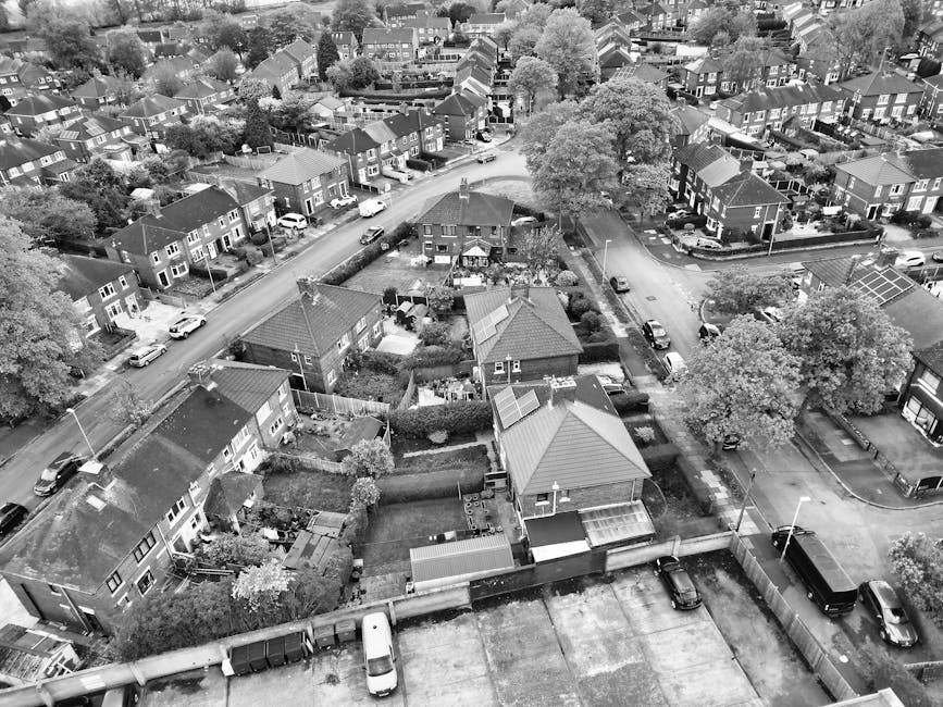 Aerial black-and-white photograph showing a residential area with numerous detached and semi-detached houses, each featuring small gardens and driveways. The houses have pitched roofs, some with solar panels, and are closely spaced along winding streets. Several cars are parked along the roads and in driveways, and a few are in motion. The image captures a section of street loading for furniture transport during home relocation, with some houses having visible outdoor packing materials like cardboard boxes and wrapped furniture, indicating ongoing moving activities. Hornchurch Man and Van's van can be seen parked on the street near the houses, with a loading process possibly underway as part of house removals or local moving services. The scene includes trees with full foliage, street lamps, and utility poles, typical of a suburban neighbourhood environment. The photograph's perspective emphasizes the tight urban layout and the logistics of moving within residential streets, relevant to the context of Havering council permits for street removals explained on the Hornchurch webpage.