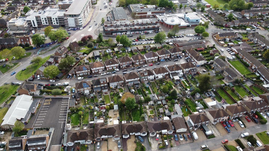 An aerial view of a residential neighbourhood in Hornchurch, showing a mix of semi-detached and terraced houses with small gardens and driveways. The houses are primarily brick-built with pitched roofs, some featuring solar panels, and are arranged in rows along narrow streets. Vehicles including cars are parked on driveways and along the street, with a few parked on the road. In the foreground, a narrow street is visible with cars parked on both sides, and behind it, the backyards of houses are enclosed with fences, some with visible garden furniture, sheds, and small sheds. To the left, a commercial building with a large parking lot is present, with several parked cars. The surroundings include trees, green spaces, and patches of lawn, with a broader urban area in the background comprising larger buildings and shopping centres. This scene is indicative of a typical home relocation area, likely captured during daylight hours, with natural lighting highlighting the details of the residential properties and moving logistics in Hornchurch. Hornchurch Man and Van may use such locations for house removals and furniture transport services, especially when planning street permit applications for moving day logistics and parking restrictions.