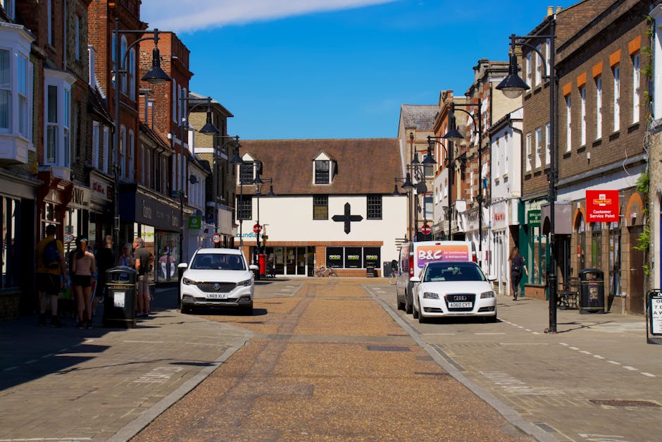 A street scene on Hornchurch High Street showing a row of multi-story retail and commercial buildings on both sides, with pedestrians walking along the pavements and a small group gathered near a bin on the left side. The street is paved with brick and has several parked vehicles, including a white van and a white Audi, positioned in the loading area close to the storefronts. The buildings feature traditional brickwork with large windows, some with signage for shops and services. Modern street lamps are mounted on black posts, and a red customer service point sign is visible on the right side near a trash bin. The sky is clear and blue, indicating daytime, and the scene appears to be set in an active commercial area suitable for home relocation and furniture transport processes, with a focus on loading and unloading activities.