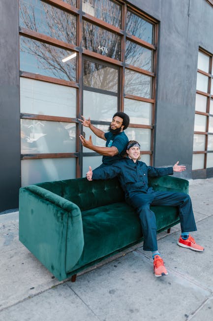 Two men are outside a commercial building with large wooden-framed windows and a grey exterior wall. One man, standing behind the green velvet sofa, is holding its backrest and smiling, while the other man is seated on the sofa with his arms outstretched and giving a thumbs-up gesture, both appearing cheerful. The scene takes place on the pavement directly outside the building, suggesting they are in the process of home relocation or furniture transport. The image includes the sofa, which is made of green velvet fabric, and shows the urban setting with natural daylight illuminating the scene. These visuals reflect packing, loading, and moving activities associated with house removals, similar to the services offered by Hornchurch Man and Van, as indicated by the webpage context.