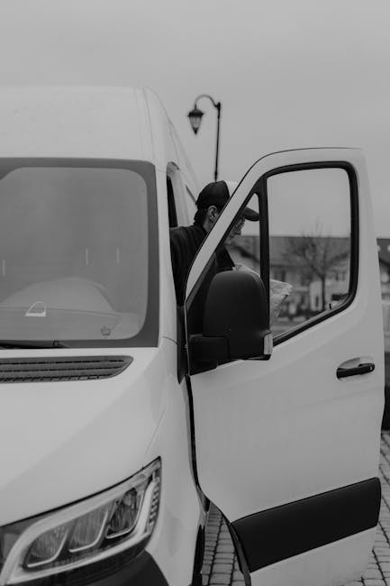 A professional mover from Hornchurch Man and Van is seen loading a large cardboard box into a white moving van parked on a paved driveway. The van’s side door is open, revealing the vehicle's interior space used for transporting household furniture and boxes. The mover is wearing dark clothing and is partially visible behind the open door, carefully carrying the box, which is sealed with packing tape. In the background, a street lamp, a few leafless trees, and residential buildings are visible under overcast sky lighting, indicating an outdoor home relocation scene. The image captures the detailed process of furniture and packing materials being prepared for a house move, highlighting the importance of efficient loading during packing and moving operations, as carried out by Hornchurch Man and Van.