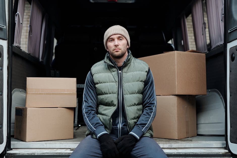 A man with a serious expression is seated on the pavement in front of an open cargo van filled with packed cardboard boxes of various sizes, some sealed with tape and others wrapped in plastic bubble wrap. The inside of the van reveals a partially visible loading area with additional packing materials like blankets and straps used for secure furniture transport. He is wearing a beige knitted beanie, a green puffer vest over a navy-blue tracksuit, and black gloves, suggesting cold weather. The background shows a residential property with a doorway and parts of the house’s exterior, indicating a home relocation scene. This image illustrates the packing and loading process involved in house removals, highlighting common steps taken in furniture transport and moving logistics, and is connected to services offered by Hornchurch Man and Van, experts in removals.
