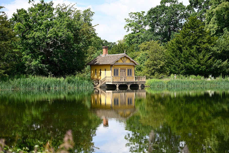 A small, yellow, wooden house with a sloped roof, situated on stilts over a calm lake surrounded by lush green trees and reeds. The house features a decorative gabled front, multiple windows with leaded glass, and a small balcony with a wooden railing extending over the water. The reflection of the house and trees can be seen clearly on the still surface of the lake. The scene is well-lit with natural daylight, emphasizing the serene environment. This setting suggests a peaceful home relocation or furniture transport scenario, in line with house removal services provided by Hornchurch Man and Van, including packing and moving logistics in the Hornchurch area.
