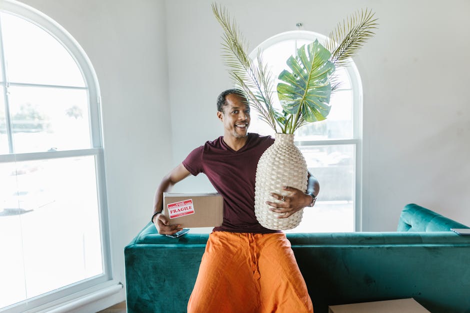 A man with dark skin and short curly hair, smiling and holding a cardboard box labeled 'Fragile' in one hand, and a large woven ceramic vase with green tropical leaves in the other, standing inside a bright room with white walls, two arched windows with natural light streaming in, and a teal sofa behind him. The setting suggests a home relocation or furniture transport scenario, with [COMPANY_NAME] providing professional removals services as part of the packing and moving process, preparing for door-to-door house and garden item transfer.