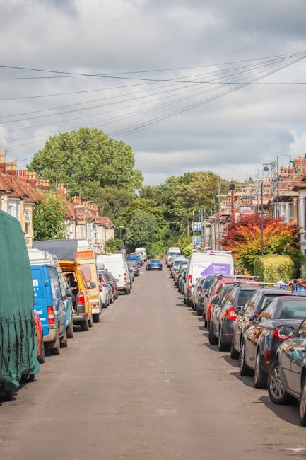 A residential street scene showing parked cars lining both sides of an asphalt road in Hornchurch, with a row of terraced houses featuring brick facades and small front gardens. In the distance, trees and greenery are visible under a cloudy sky. The street appears quiet, and there are overhead power lines running across the scene. This setting reflects a typical suburban neighbourhood relevant for household relocations and local house removals, as offered by Hornchurch Man and Van. The image emphasizes the importance of careful parking and logistics considerations during home relocation or furniture transport, illustrating the environment where a professional removals service might operate within the local streets of Hornchurch, aligning with the RM11 moving guide.