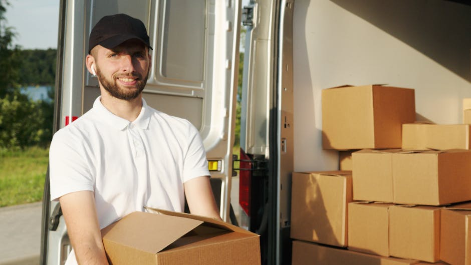 A man wearing a white polo shirt and a black cap is smiling and holding a cardboard box during a home relocation process. He is standing next to an open van with a large cargo space, which is partially visible behind him. Inside the van, there are several neatly stacked cardboard boxes of various sizes, some wrapped in plastic or placed directly on each other. The loading area is outdoors on a paved surface, with a green landscape and trees in the background, illuminated by natural daylight. This scene captures the loading process involved in furniture transport and packing for house removals, with the man's position indicating active engagement in the moving logistics. The setting reflects professional moving and packing services provided by Hornchurch Man and Van as part of their residential moving solutions, shown in the context of a local street environment, supporting the relevance of the RM11 moving guide for Hornchurch.