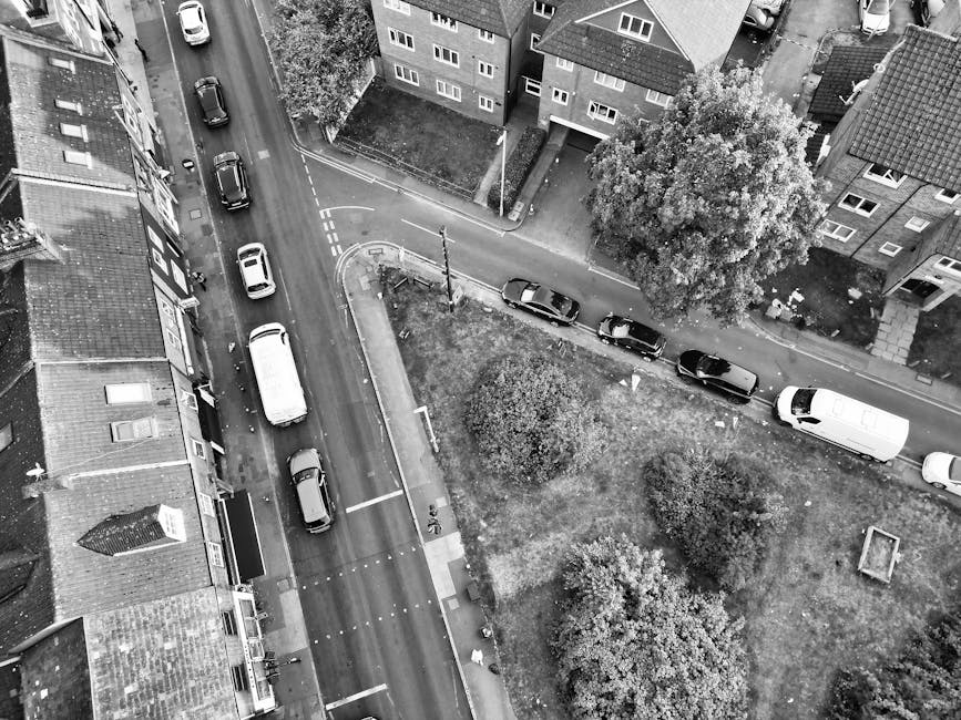 A residential street scene showing parked cars lining both sides of an asphalt road in Hornchurch, with a row of terraced houses featuring brick facades and small front gardens. In the distance, trees and greenery are visible under a cloudy sky. The street appears quiet, and there are overhead power lines running across the scene. This setting reflects a typical suburban neighbourhood relevant for household relocations and local house removals, as offered by Hornchurch Man and Van. The image emphasizes the importance of careful parking and logistics considerations during home relocation or furniture transport, illustrating the environment where a professional removals service might operate within the local streets of Hornchurch, aligning with the RM11 moving guide.